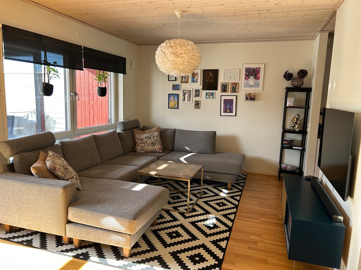 A spacious living area features a comfortable sectional sofa adorned with decorative pillows. A small table is positioned in front of it, complemented by a patterned rug. Natural light floods the room through large windows, which are covered with stylish blinds, illuminating the surrounding art and decorative elements.