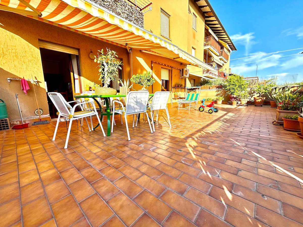 A spacious terrace is presented, featuring a dining table surrounded by chairs and potted plants. Sunlight illuminates the area, highlighting the terracotta tiled flooring. Colorful striped awnings provide shade, while a small child's tricycle adds a playful touch to the scene.