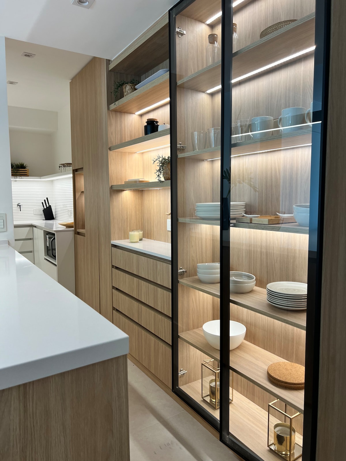 A modern kitchen area is displayed with wooden cabinetry and glass shelving. Soft lighting highlights a variety of dishware and decorative items. A countertop is visible on the left, enhancing the clean and organized appearance of the space, which fosters a sense of functionality.