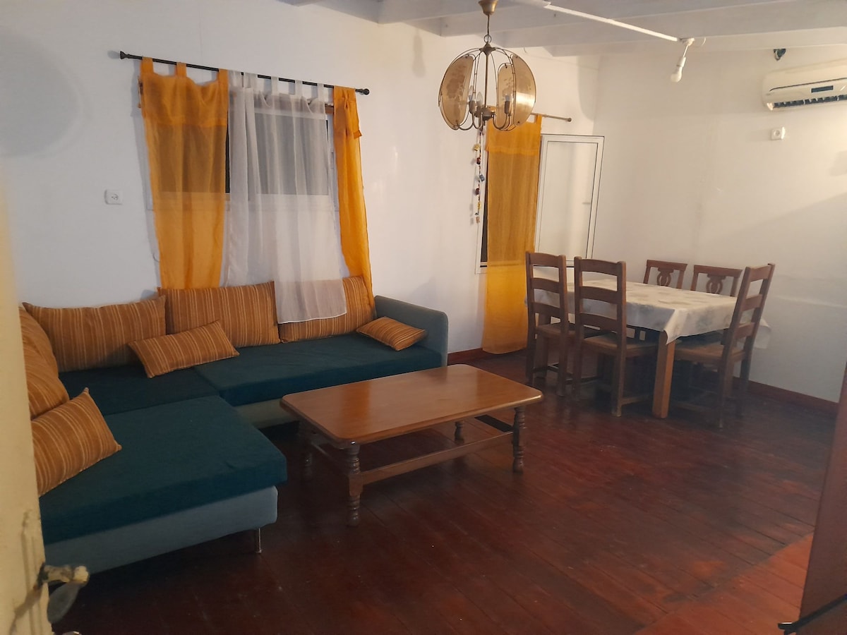 A living area features a spacious sectional sofa with striped cushions alongside a wooden coffee table. A dining table with wooden chairs occupies one corner, while bright orange curtains frame the windows, allowing natural light to brighten the room.