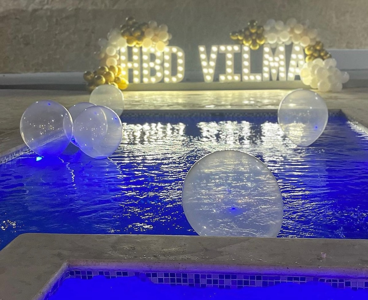A swimming pool is illuminated with a soft blue light, creating a calming atmosphere. Transparent beach balls float on the water's surface. A festive display features the text 'HBD VILMA' adorned with decorative balloons in gold and white, enhancing the celebratory vibe.