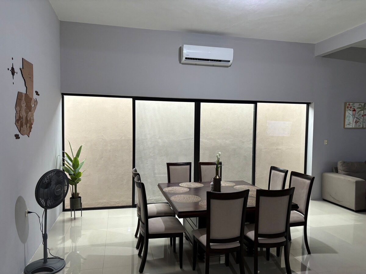 A spacious dining area is presented, featuring a large table with eight chairs. The room is bathed in natural light from expansive windows that overlook a serene outdoor space. A small potted plant adds a touch of greenery to the setting.