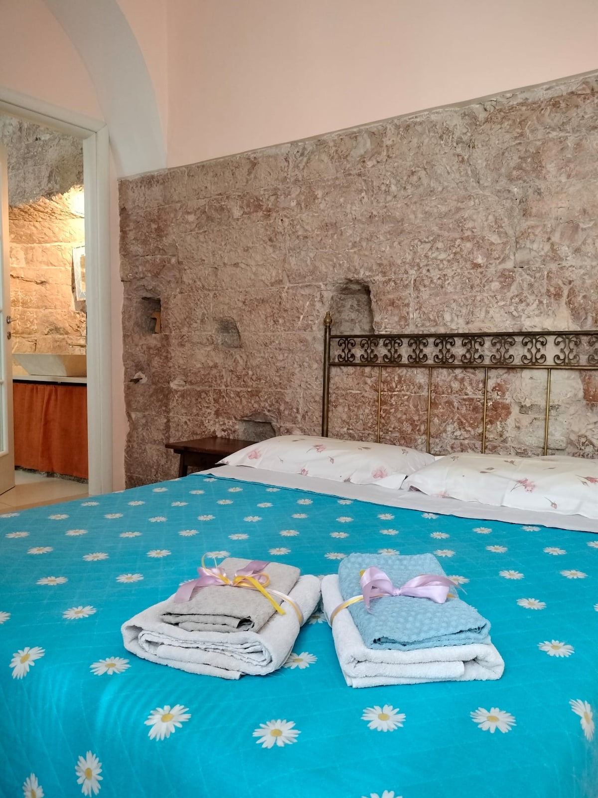 A spacious sleeping area is marked by a bright blue bedspread adorned with floral patterns. Neatly folded towels are placed at the foot of the bed. The rustic wall, featuring original ancient stone, complements the inviting atmosphere of the room.