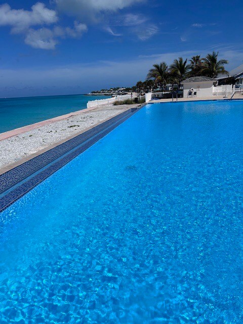 A crystal-clear swimming pool reflects the bright blue sky, with the ocean visible in the background. Several palm trees line the shore, and a sandy beach extends alongside the pool, creating a serene atmosphere.