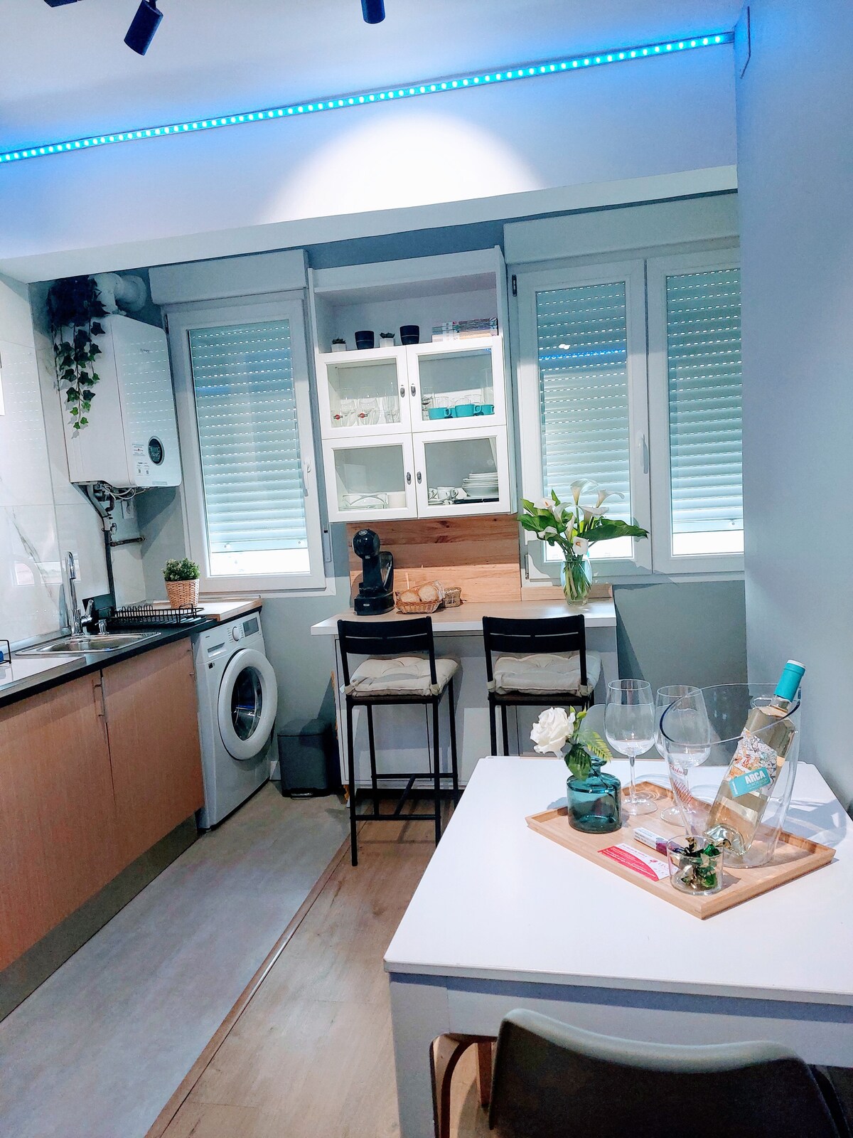 A compact kitchen area is featured, showcasing stainless steel appliances, including a washing machine and coffee maker. Natural light streams through multiple windows, illuminating a small dining table set with glasses and a bottle of wine, creating a functional and inviting space.