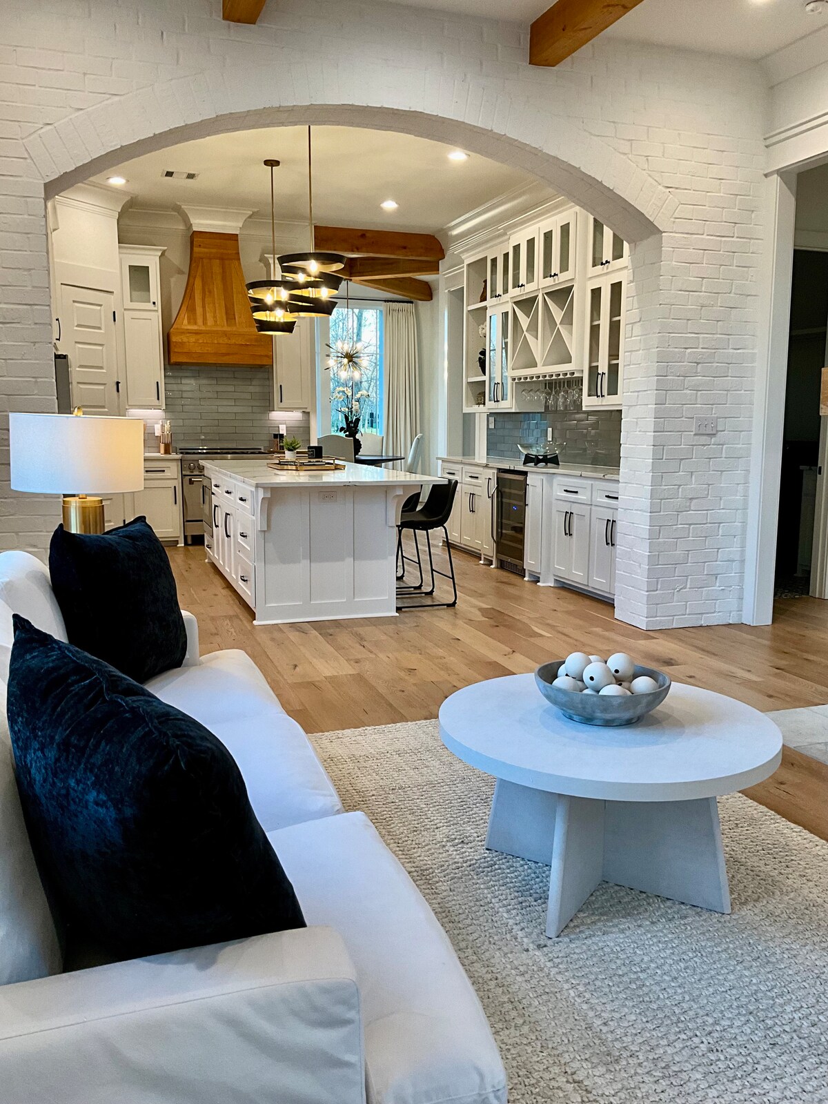 A modern kitchen and living area are showcased, featuring a spacious island with bar seating. Light-colored cabinetry contrasts with the wooden ceiling beams. A cozy seating arrangement is present, accented by dark pillows and a decorative bowl on a low round table.