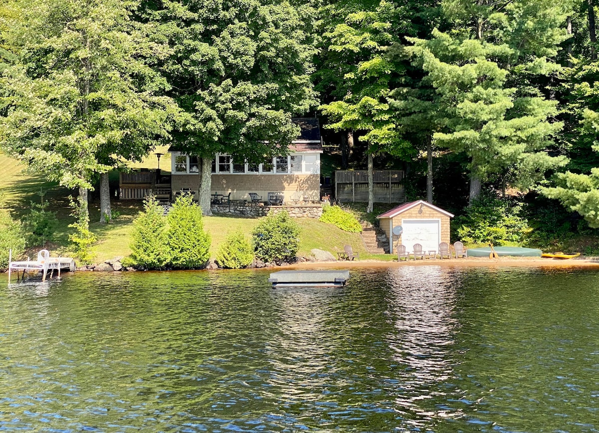 A rustic cabin is visible along the edge of the Racquette River, surrounded by lush trees. Seating areas are arranged on a sandy beach, with a dock extending over the water. The calm, reflective surface of the river adds to the serene setting.