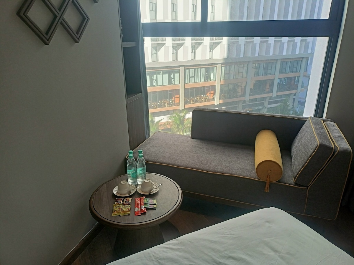 A lounge area includes a gray chaise with a yellow cushion beside a round table. Two bottles of water and two plates with cups are arranged on the table, with snacks positioned nearby. Large windows provide a view of the surrounding architecture.