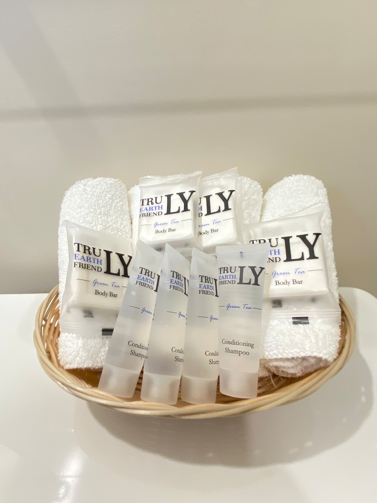 A woven basket holds neatly arranged, white terry towels alongside complimentary travel-sized toiletries. The items include conditioning shampoo, body bar, and body wash, all labeled for easy identification. The presentation is organized and contributes to a tidy bathroom setting.