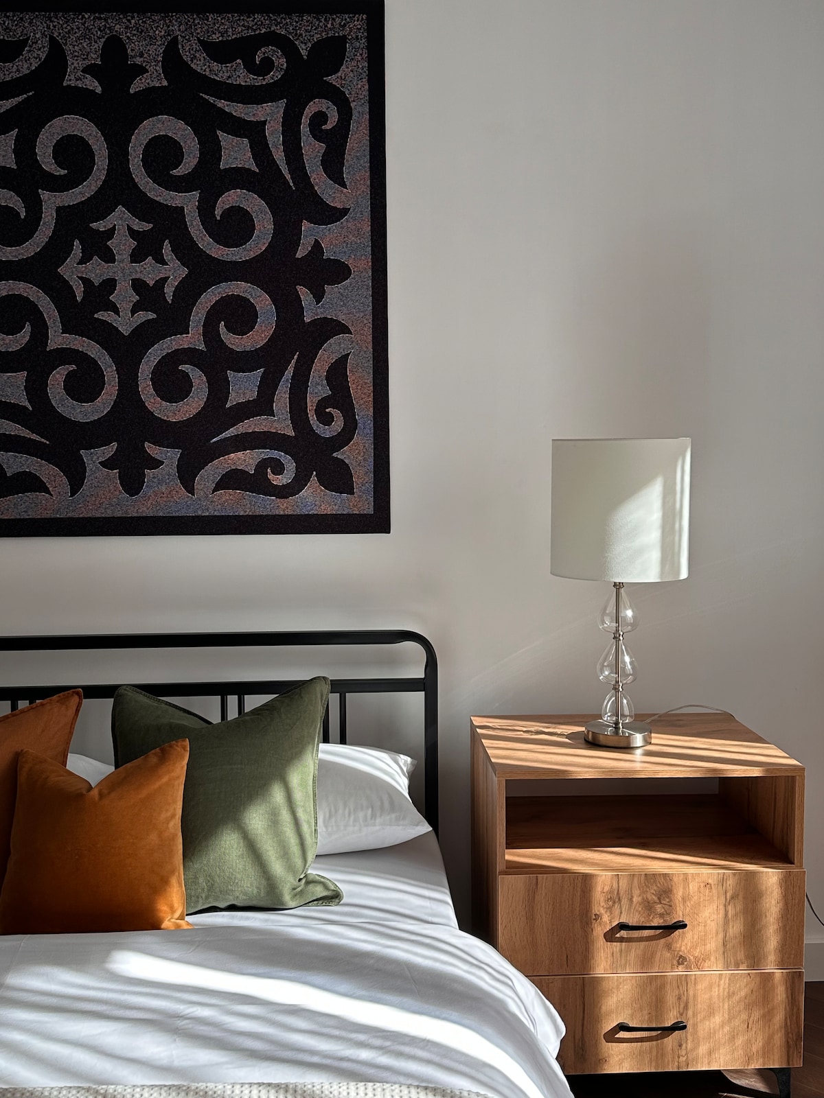 A modern bedroom setting is featured, showcasing a sleek black bed with decorative cushions in green and orange. A wooden nightstand with two drawers is positioned beside the bed, topped by a stylish lamp that casts soft shadows.