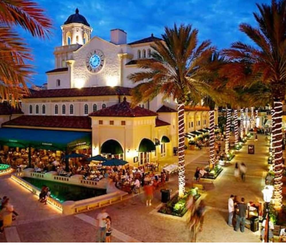 An outdoor dining area is featured, surrounded by palm trees and lit by twinkling lights. The historic building serves as a backdrop, showcasing an ornate clock tower. A lively atmosphere is created by people enjoying the space, with seating areas extending along the plaza.
