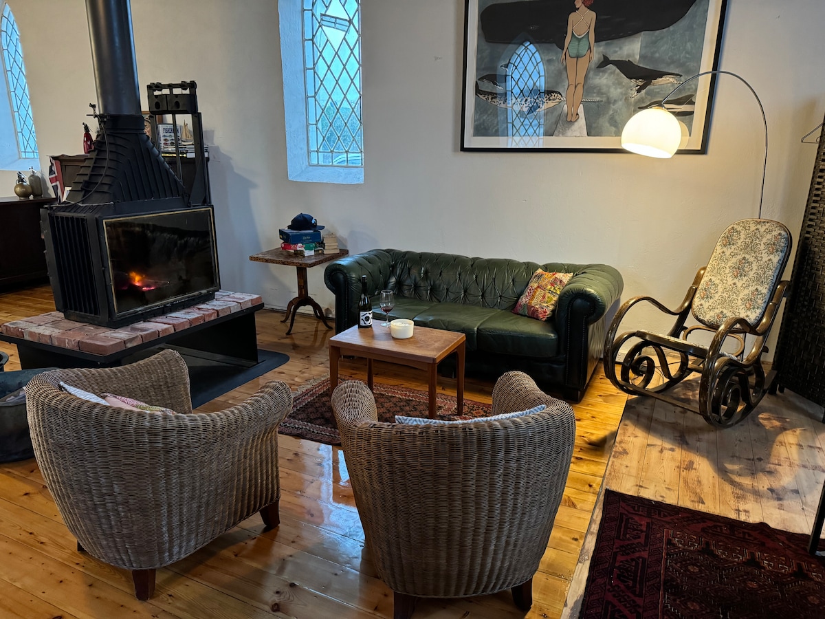 A cozy living area features a double-sided fireplace surrounded by comfortable seating, including two woven chairs and a vintage rocking chair. A wooden coffee table sits in the center, complemented by a colorful cushion, with light streaming in through arched windows.