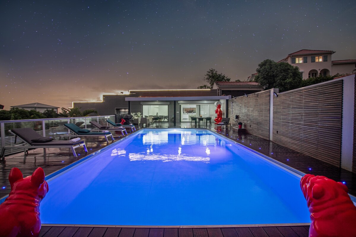 A modern, illuminated swimming pool is situated within a spacious outdoor area, surrounded by sleek lounging chairs. Two vibrant red sculptures flank the poolside, complementing the sophisticated atmosphere. The villa's exterior is visible in the background, featuring expansive glass doors that invite great views of the outdoor space.