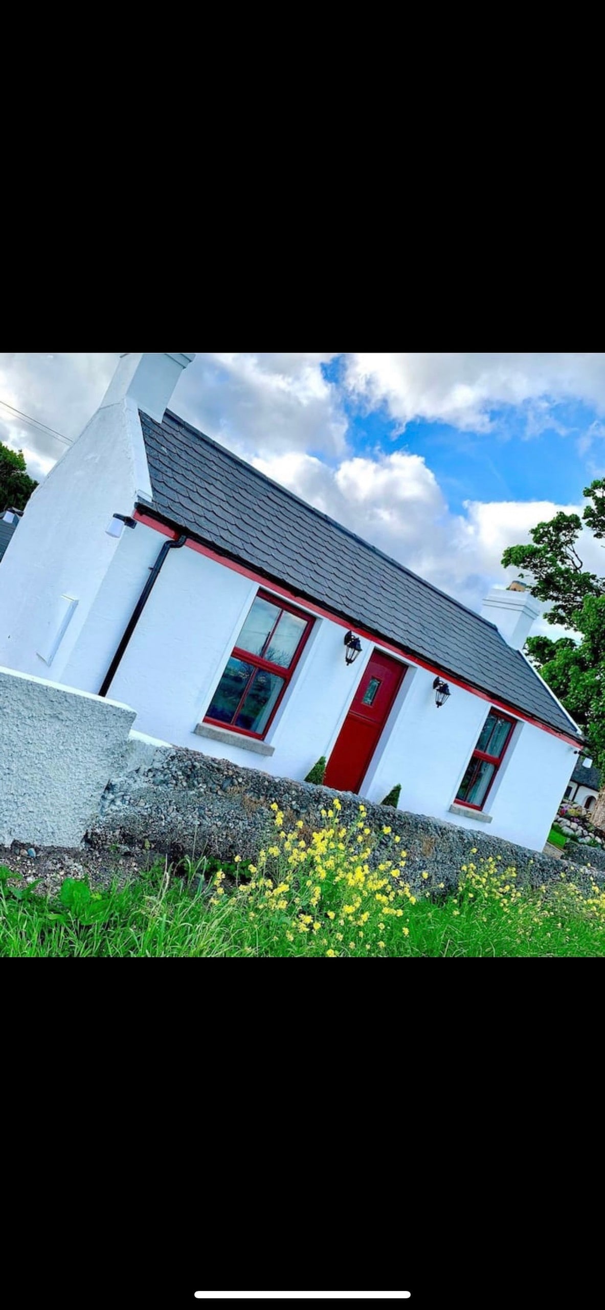 A charming white cottage is set against a vibrant landscape, featuring a red front door and matching window frames. Lush greenery and flowering plants surround the property, while a stone wall adds rustic character. Soft clouds pass overhead, enhancing the serene environment.