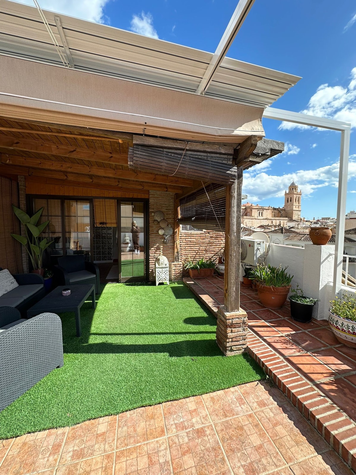 An outdoor terrace features synthetic grass and a wooden pergola, providing a shaded area. Potted plants are positioned around the space, enhancing the serene atmosphere. City views are visible beyond the railing, with a historical tower visible in the background under a clear blue sky.