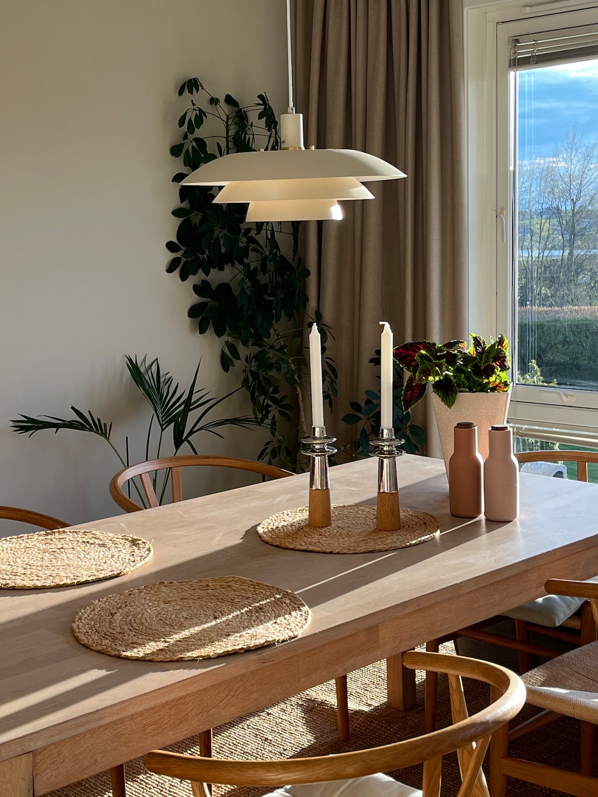 A dining area is presented with a wooden table surrounded by light-colored chairs. A stylish lamp hangs overhead, illuminating the space. Two candles in silver holders are positioned on the table, alongside woven placemats. A vase with flowers is placed by the window, framed by curtains.