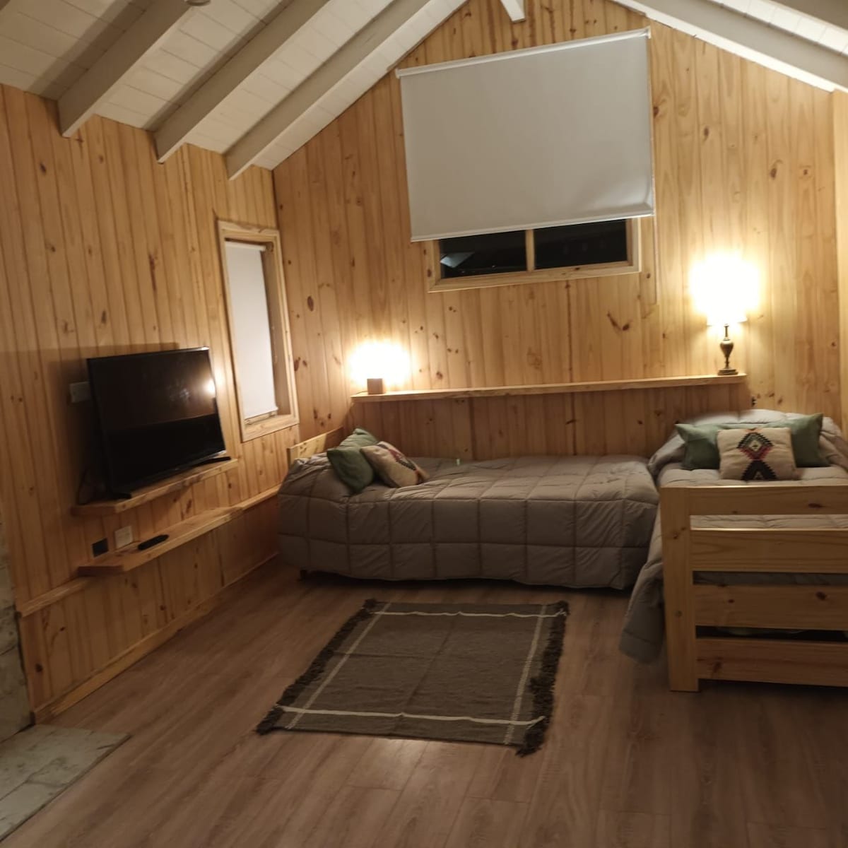 The interior space features wooden walls and ceilings, contributing to a cozy ambiance. Two beds are arranged in the room, with a small TV mounted on the wall. A woven rug is placed on the light-colored floor, and soft lighting enhances the overall warmth.