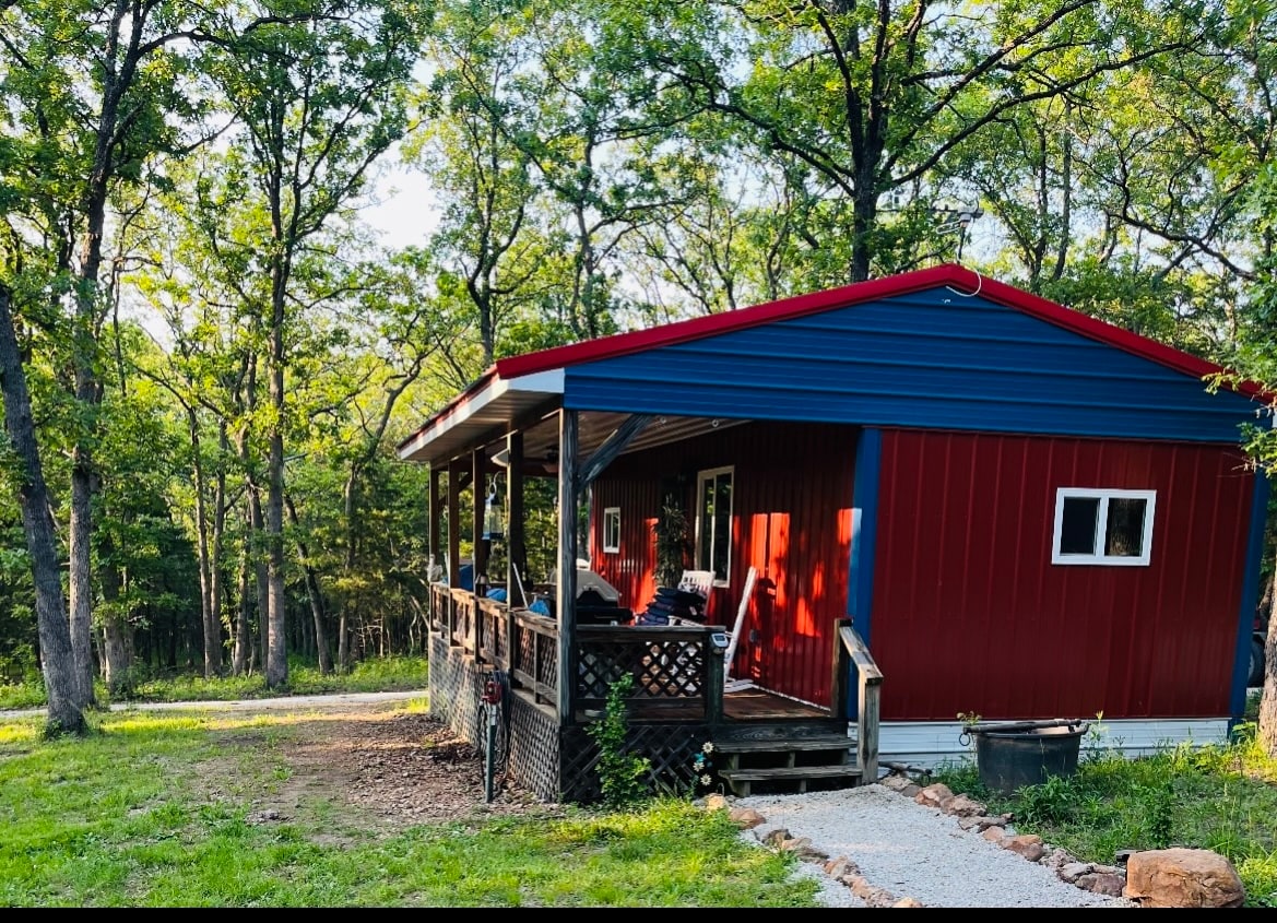A modern cabin with red and blue exterior is situated among trees. The spacious front porch features a railing and is partially shaded by the overhang. A gravel path leads to the entrance, surrounded by grassy areas and woodland.