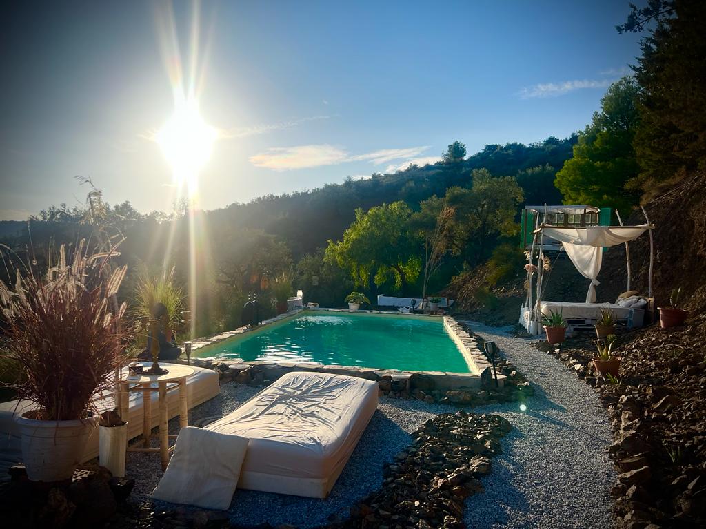 A sunlit pool area features a large turquoise swimming pool surrounded by natural stones. Comfortable loungers are positioned nearby, and a shaded seating area is visible to the side, framed by greenery and decorations. The landscape showcases gentle hills in the background.