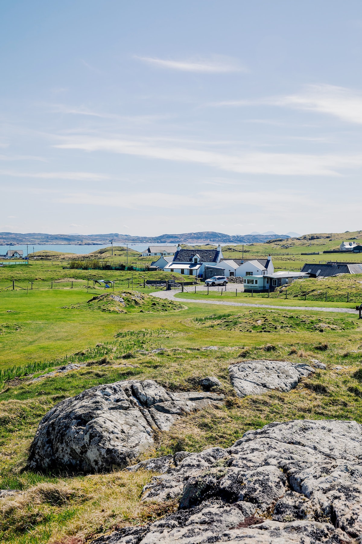 A scenic view of a rural landscape showcases a collection of modernized croft houses nestled among green fields. Rolling hills and rocky outcrops are visible, with a distant view of the coastline under a clear blue sky.