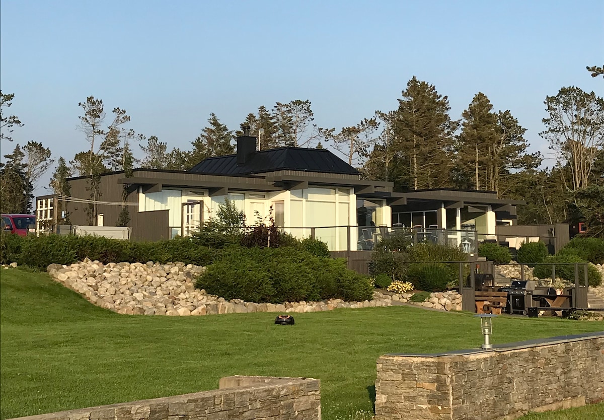 A modern retreat is showcased in this image, featuring large glass windows and a sleek black roof. The surrounding landscape includes well-maintained greenery and rocky stone borders. Towering trees are visible in the background, contributing to a calming and natural environment.