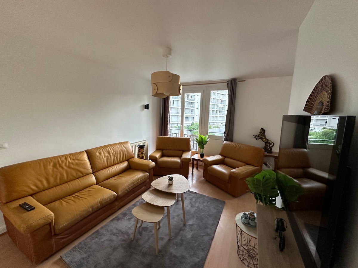 A cozy living area features brown leather sofas arranged around a light wooden coffee table. A small potted plant sits on the table, and a textured rug covers the floor. Natural light enters through large windows, with sheer curtains providing a soft touch.
