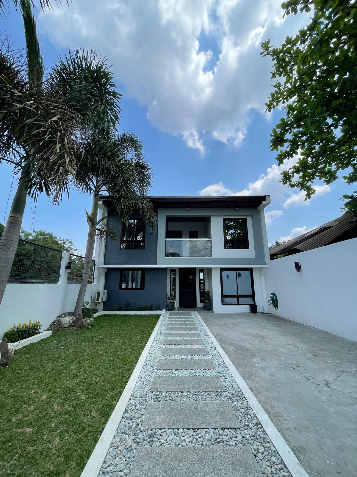 The exterior of a modern two-story house is presented, featuring a well-maintained pathway lined with gravel and grass. Lush palm trees are visible in the front yard, and large windows reflect natural light. A balcony is seen on the upper floor, enhancing the overall contemporary design.