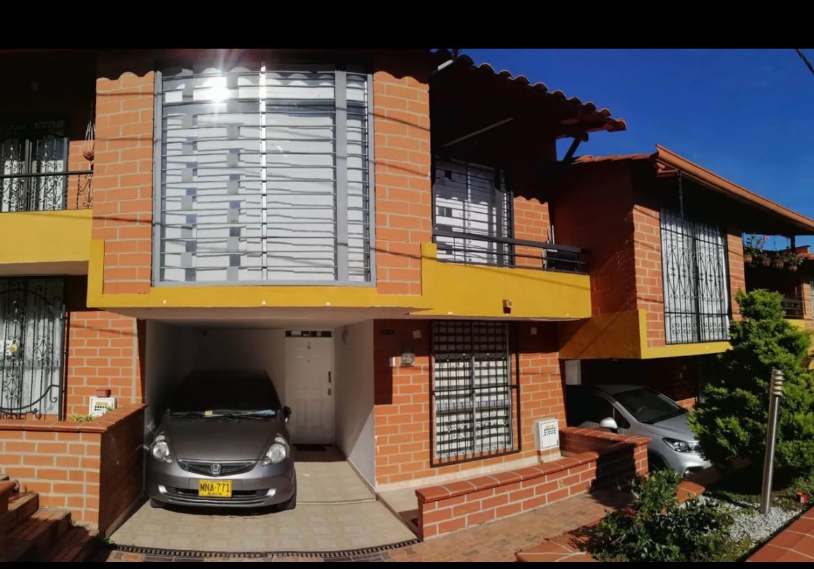 The image showcases a two-story, brick house featuring large, horizontal window slats that allow natural light to enter. A vehicle is parked in the carport, which is accessed by a driveway lined with decorative stones. Bright blue sky enhances the welcoming appearance of the home.