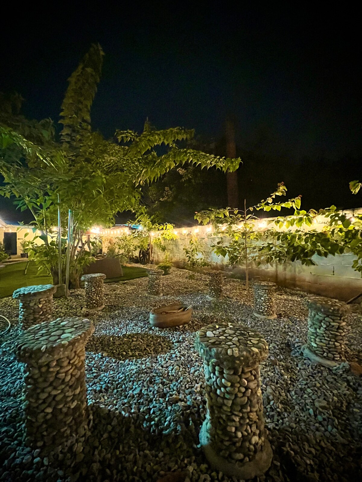 A peaceful outdoor space is surrounded by stone pillars arranged in a circular formation, offering seating. Soft lighting illuminates the area, highlighting the textured pebble ground and surrounding greenery, creating an inviting ambiance for gatherings.