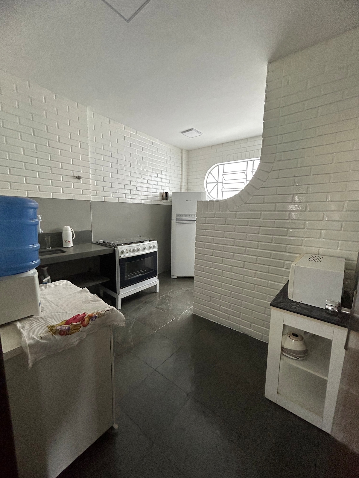 A functional kitchen space features white brick walls and dark countertops. A gas stove is positioned against one wall, with a refrigerator adjacent to it. A water dispenser is visible on the counter, complemented by a microwave on a nearby shelf. Natural light enters through a window.
