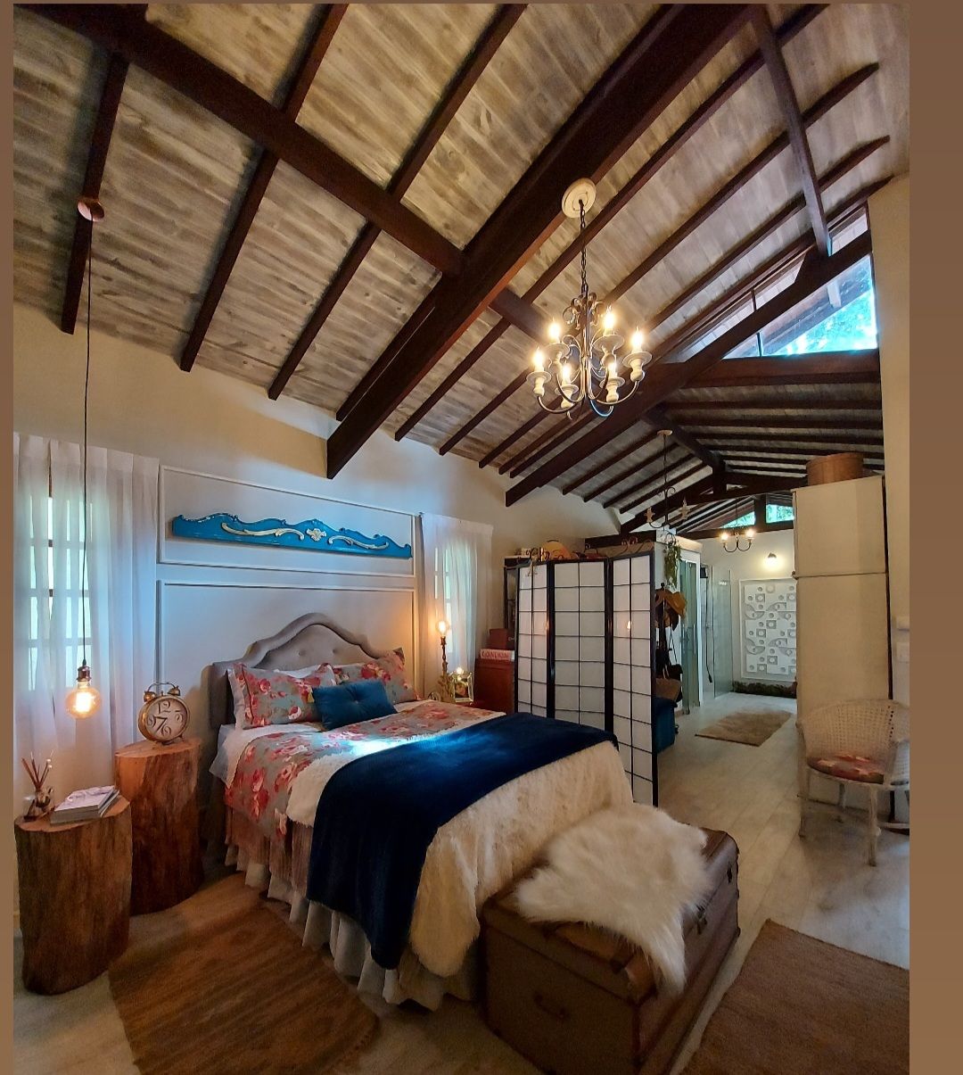 A spacious bedroom features a high, wooden beamed ceiling and a comfortable bed adorned with colorful bedding and decorative pillows. A two-sided screen separates the bedroom area from an adjoining space. Natural light filters through sheer curtains, highlighting wooden side tables and a decorative chandelier.