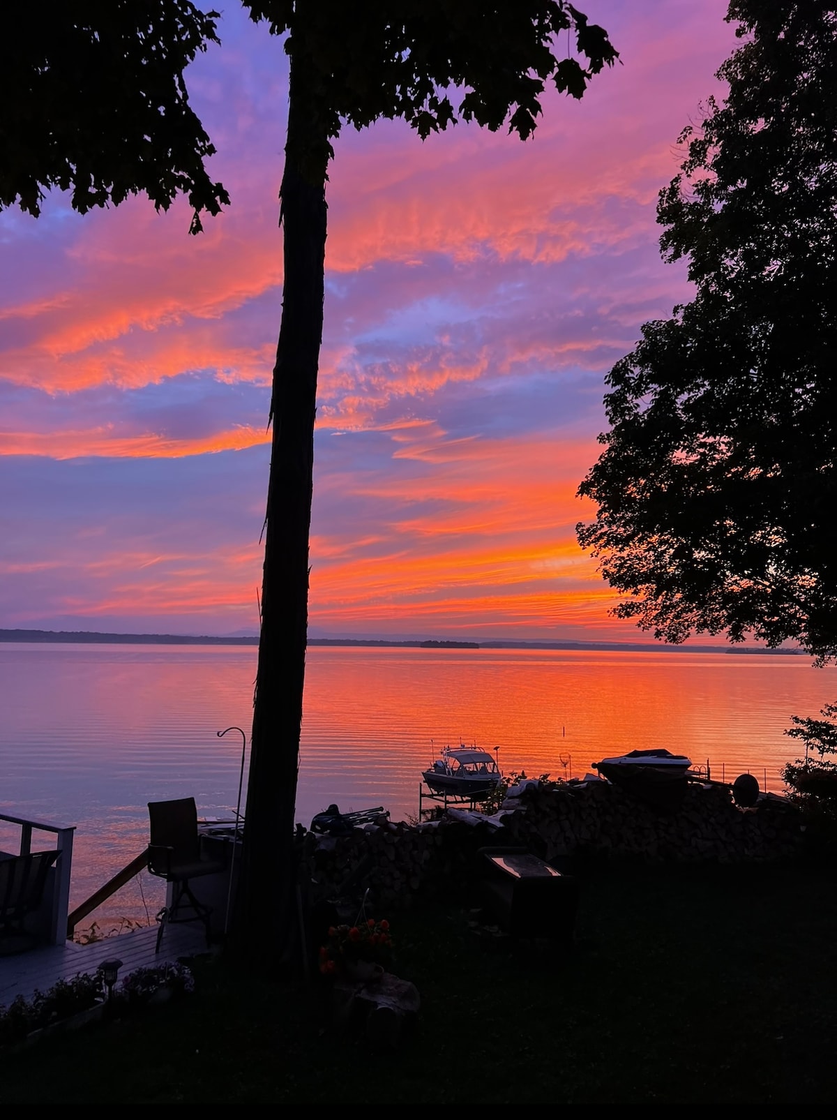 A tranquil waterfront scene captures a stunning sunset over the water, showcasing vibrant hues of orange and purple in the sky. Silhouettes of trees frame the view, while boats are docked peacefully along the shore, contributing to the serene atmosphere.