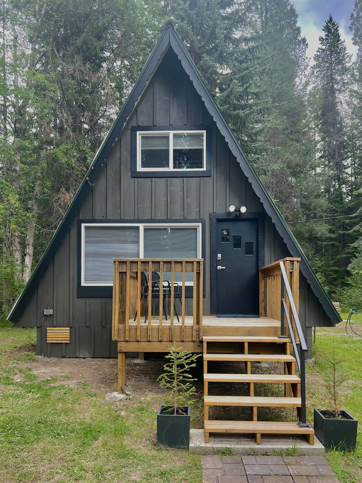 The exterior of a rustic-modern A-frame cabin is showcased, featuring dark wooden siding and large windows beneath a pointed roof. A wooden staircase leads to the entrance, accompanied by a small outdoor seating area and greenery surrounding the cabin.
