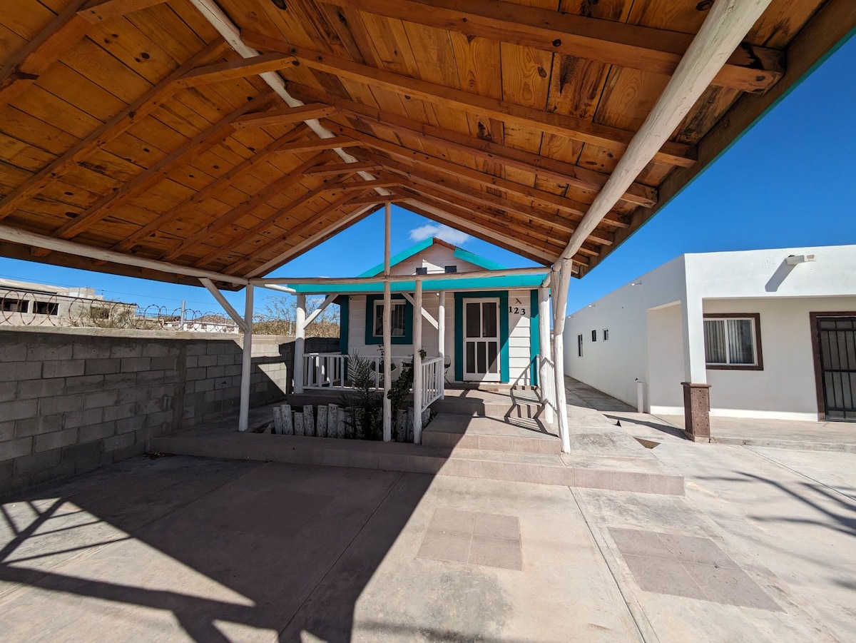 The exterior features a covered parking area with a wooden roof and a concrete driveway leading to the entrance. A welcoming porch is adorned with white railings, while the house displays a light blue facade against a clear sky.
