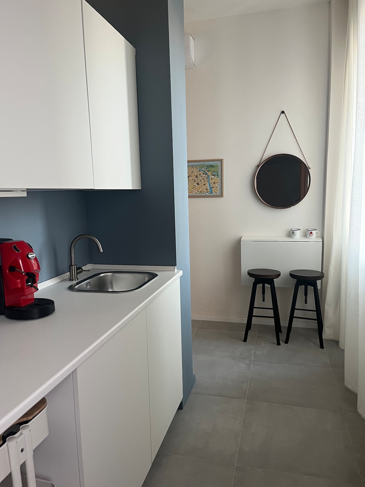 The kitchenette features modern cabinetry in white, equipped with a small sink and a coffee machine. A round mirror hangs on the wall opposite a compact dining area with two black stools and a small table, illuminated by natural light from the adjacent window.