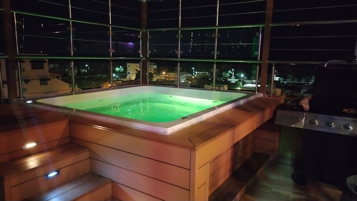 A spacious hot tub is surrounded by wooden decking, with colorful lights casting a gentle glow on the water. Glass panels provide an unobstructed view of the city skyline at night, enhancing the serene atmosphere.