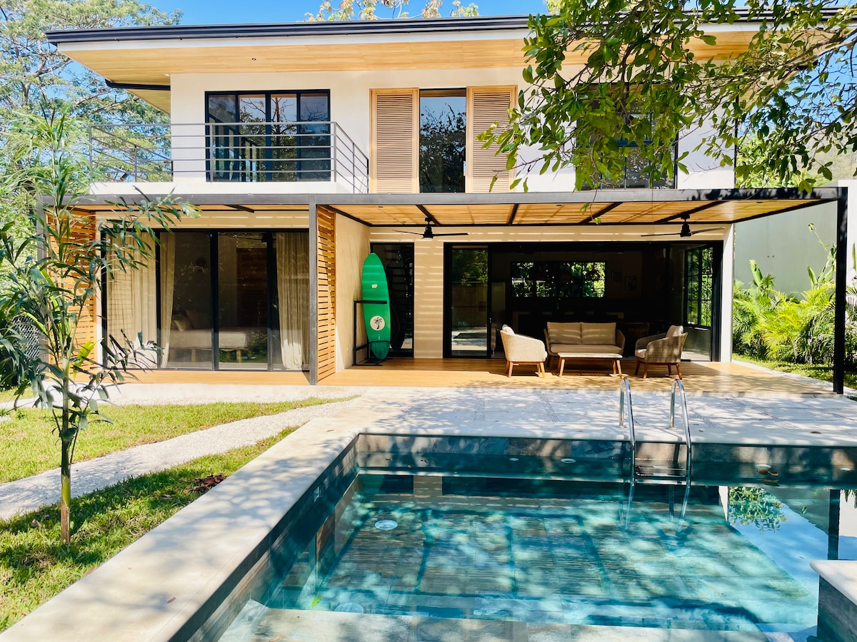 A modern house is shown with a spacious terrace overlooking a private swimming pool. Large glass doors provide a view into the living area, and outdoor seating is positioned under the wooden overhang. Lush greenery and tropical plants frame the serene setting.