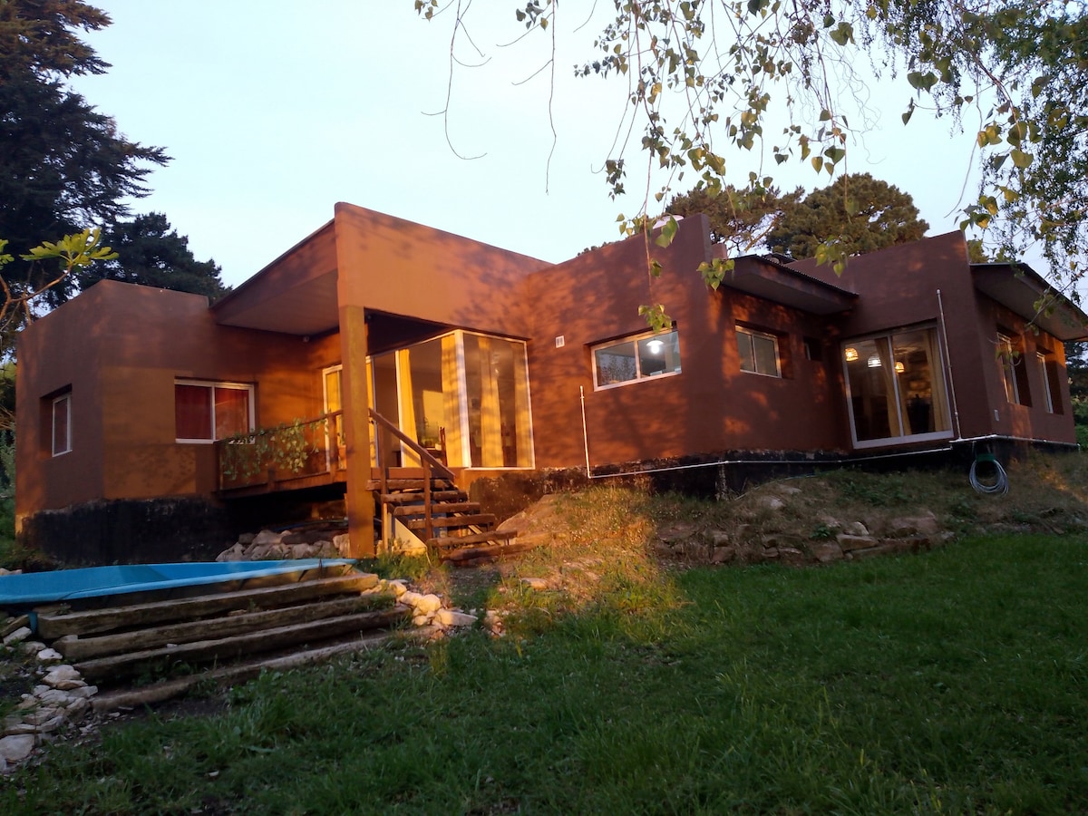 A modern structure is surrounded by green grass and trees, featuring large glass windows that allow natural light to fill the interiors. A set of wooden stairs leads to the entrance, while a small water feature is situated nearby, complementing the outdoor setting.