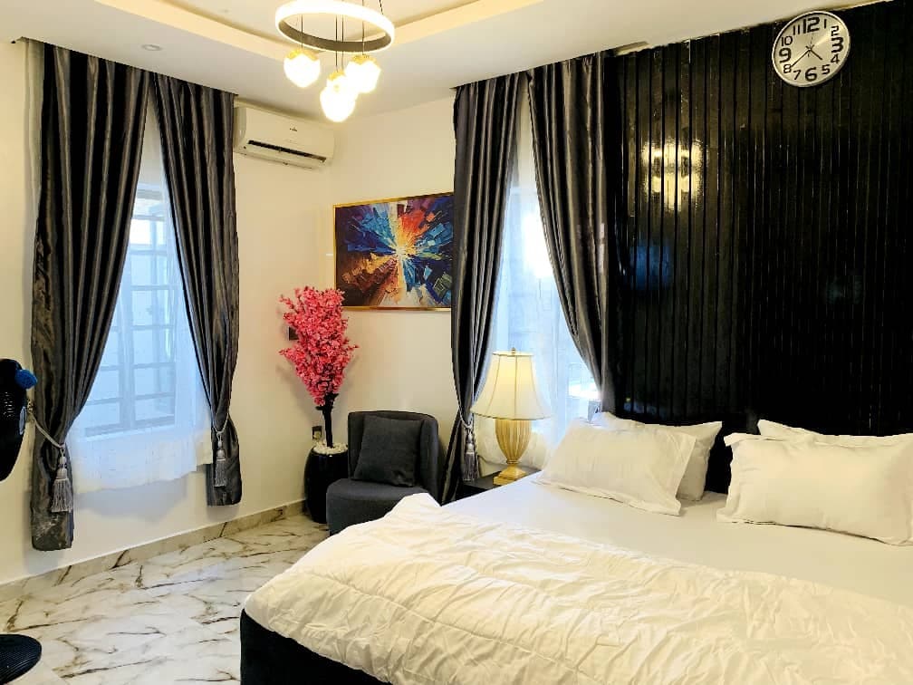 A well-lit room features a spacious bed dressed in crisp linens. Dark curtains frame the windows, while a piece of abstract art adds color to the wall. A modern lamp and a decorative plant enhance the cozy atmosphere, and a clock is mounted on the wall.
