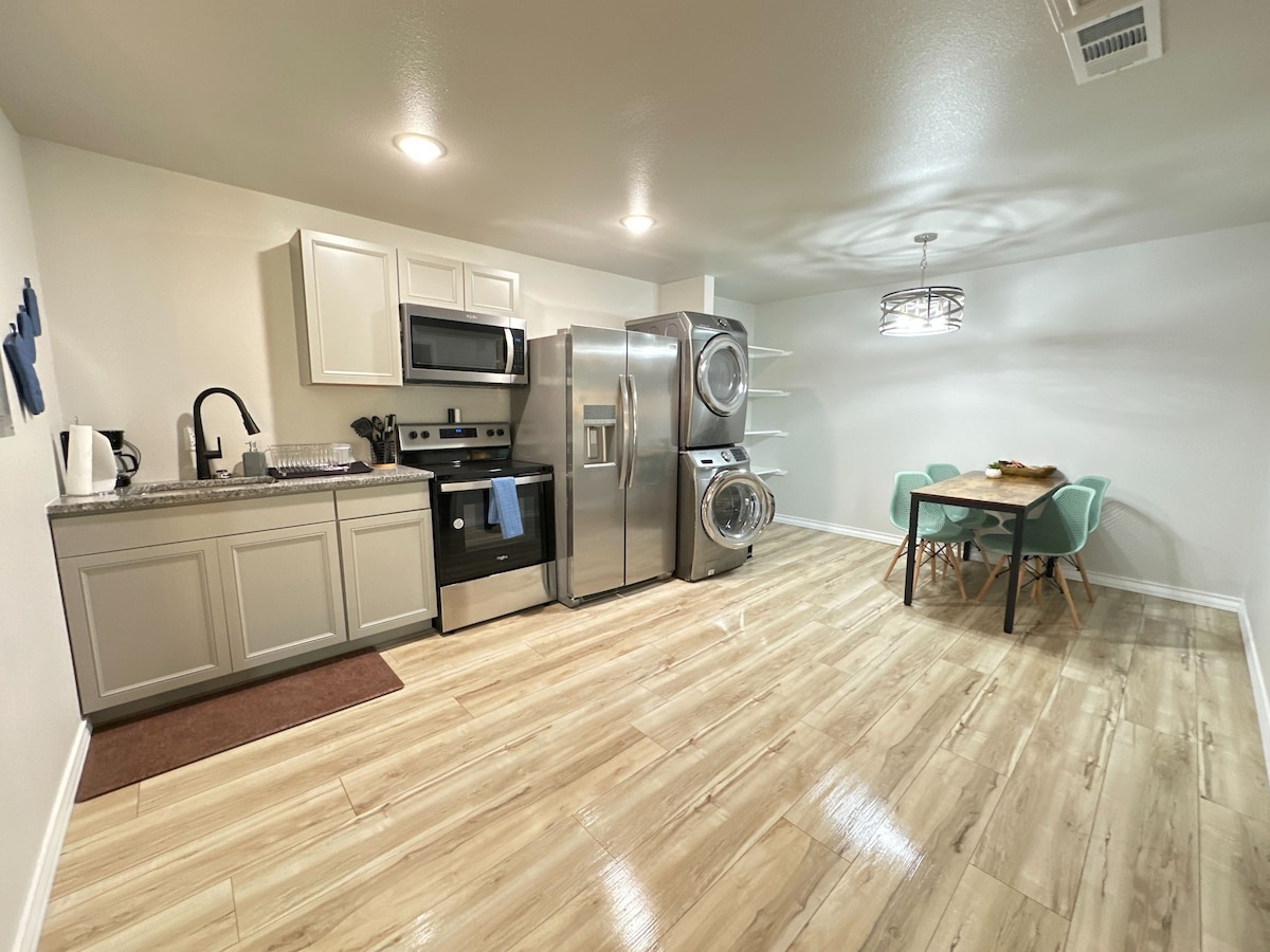 A modern kitchen is equipped with stainless steel appliances, including a refrigerator and a washer-dryer unit. Light-colored cabinetry complements the granite countertop. A dining area features a wooden table surrounded by four mint-green chairs, set against a backdrop of warm, light-colored flooring.