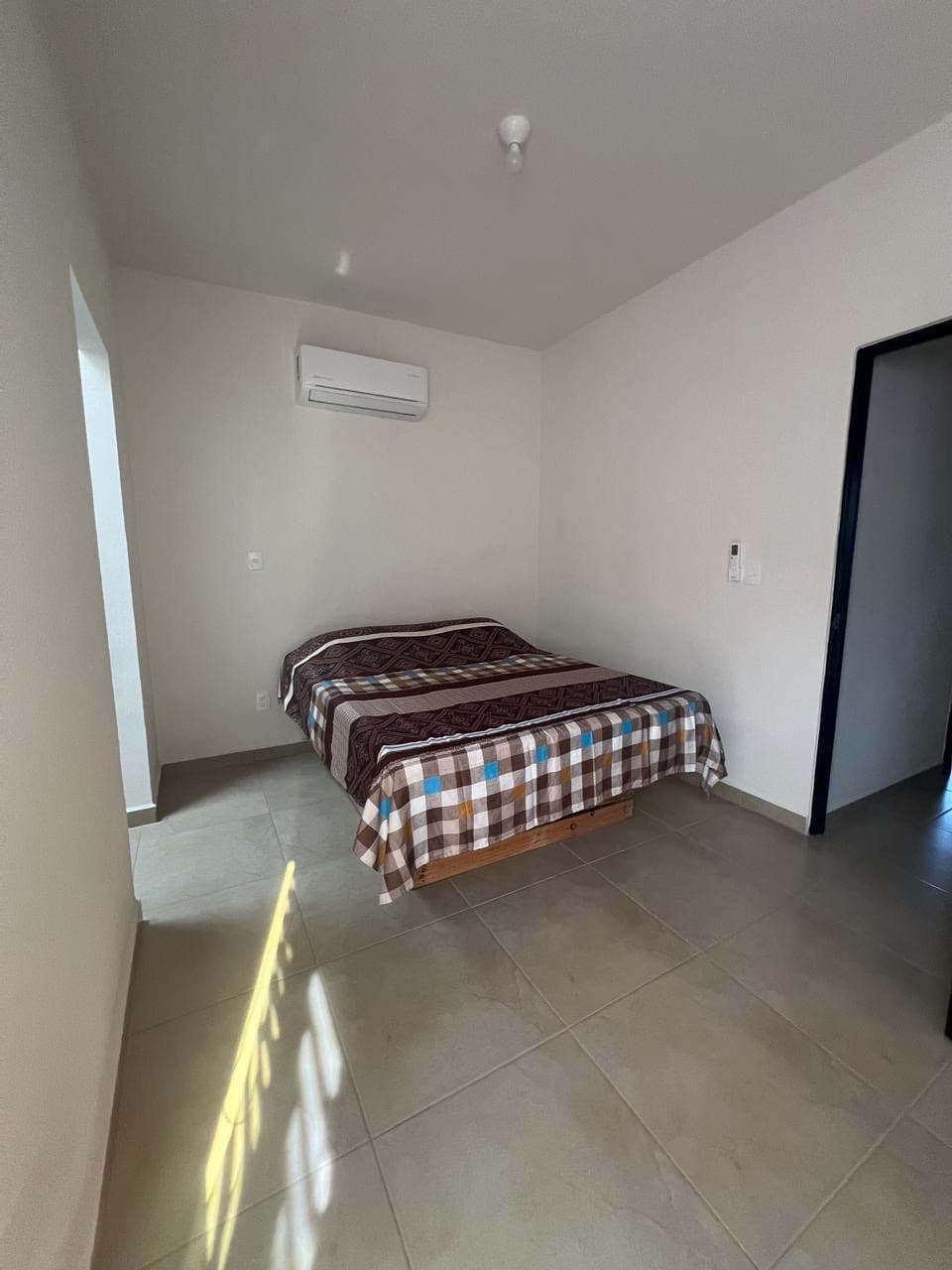 A simple and modern bedroom is shown, featuring a spacious bed covered with a striped and checkered blanket. Natural light filters in, highlighting the neutral-toned walls and floor tiles. An air conditioning unit is mounted on the wall for comfort.