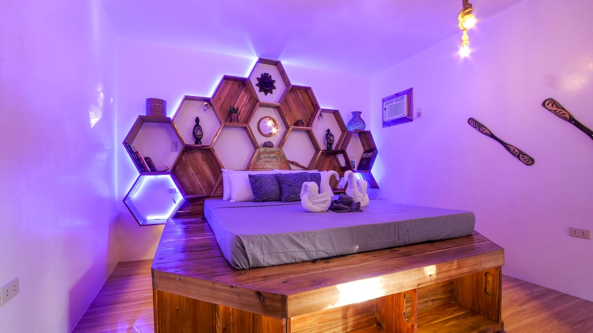 A unique sleeping area features a hexagonal wooden shelf display, adorned with decorative items and ambient lighting. A spacious bed is centered in the room, topped with crisp linens. The warm wood tones and soft lighting contribute to a cozy environment.