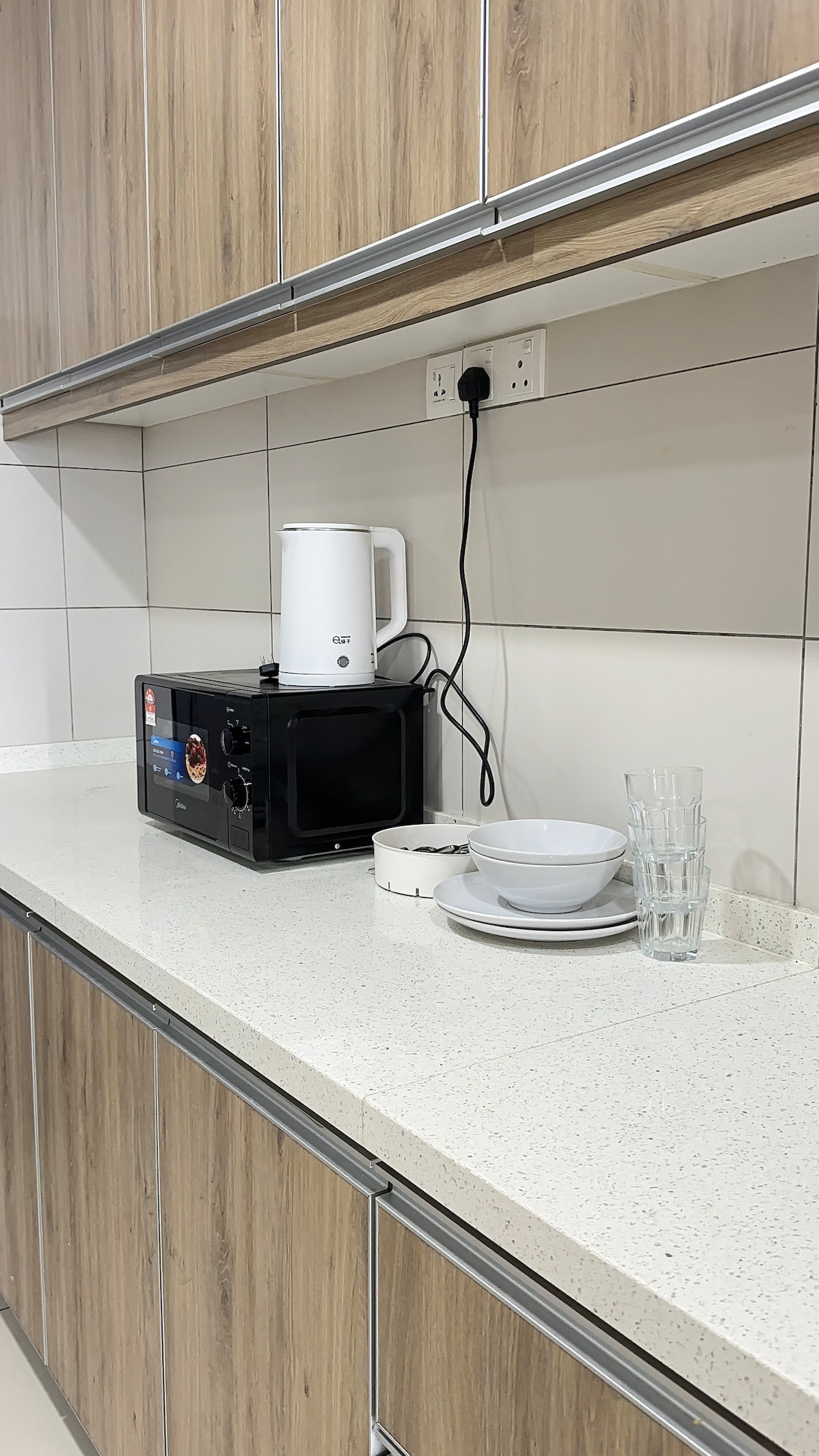 A kitchen counter features a microwave and kettle alongside a set of white bowls. Two clear glasses are positioned next to the bowls, all arranged neatly against a backdrop of wood and cream-colored cabinets.