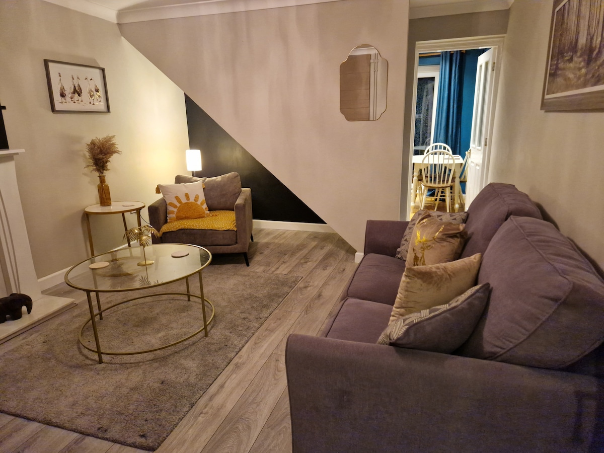 A cozy lounge area features a grey sofa and armchair, arranged around a circular coffee table. A plush area rug covers the floor. The walls are decorated with minimalist art, and a stylish lamp provides soft illumination. A dining area can be seen through an open doorway.