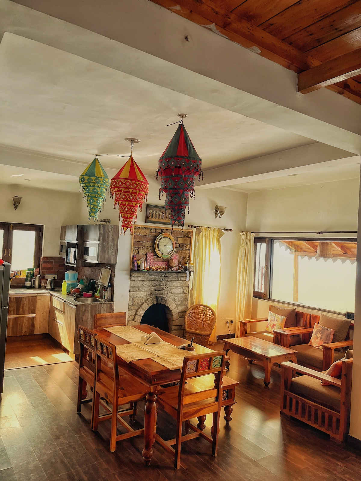 An open dining area features a large wooden table surrounded by sturdy chairs. Colorful hanging lanterns add character to the space, while a stone fireplace is positioned against one wall. Windows allow natural light to brighten the room, complemented by warm wooden accents.