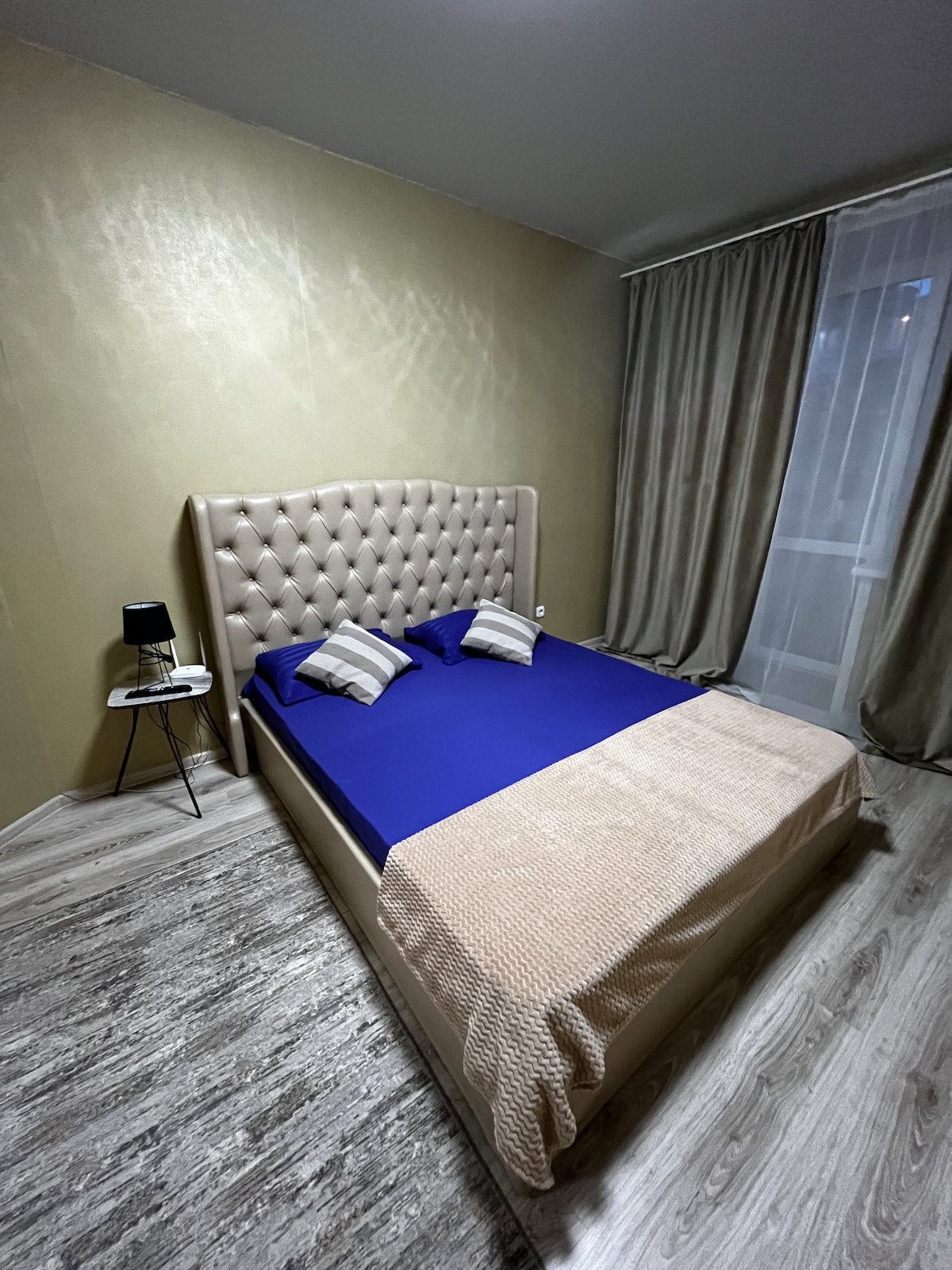 A cozy sleeping area features a spacious bed with a tufted headboard and blue bedding. Two decorative pillows are positioned on the bed. Light-colored curtains frame the window, and a small bedside table with a lamp is placed next to the bed.