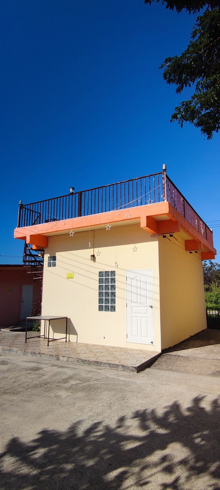 The exterior of a brightly painted two-story building is shown, featuring a yellow facade with orange accents on the rooftop. A staircase is visible on the side, leading to a rooftop area surrounded by a metal railing. Clear blue skies complement the overall setting.