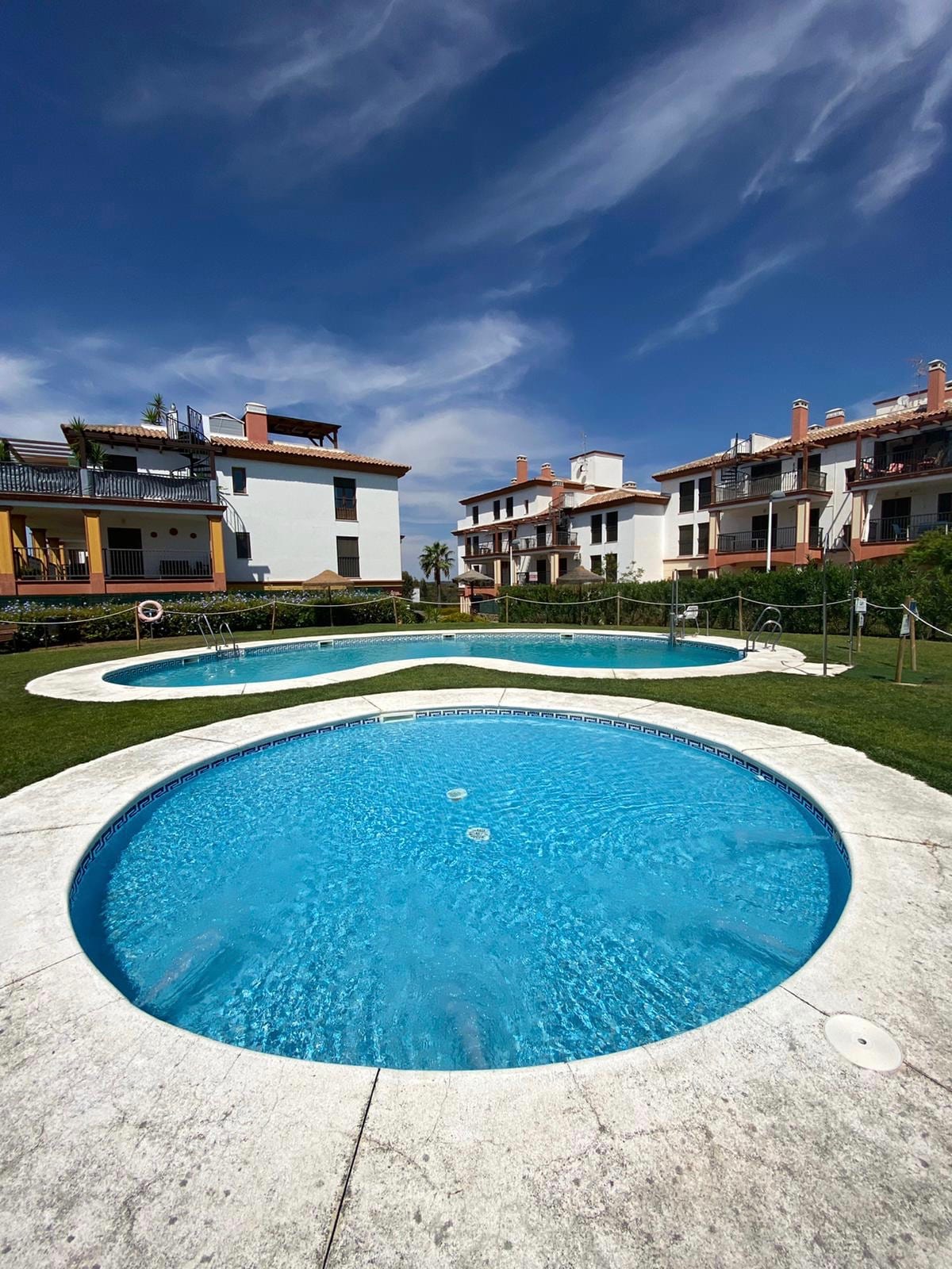 Two circular swimming pools are surrounded by a grassy area, with several residential buildings in the background. The crystal-clear water reflects the blue sky, while sun loungers are positioned along the poolside for relaxation.