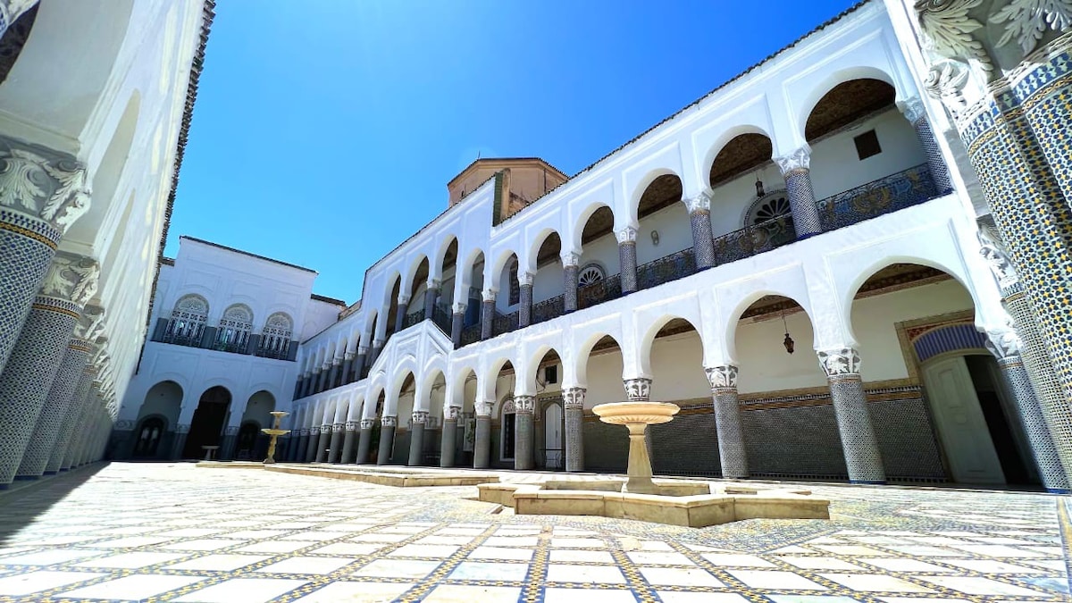 An expansive courtyard is surrounded by elegant arches and intricately tiled balconies. A central fountain adds a calming feature to the sunlit space, where vibrant mosaic patterns cover the ground, creating a harmonious connection between architecture and design.