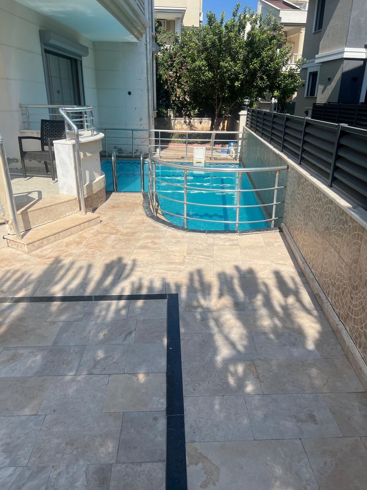 The image shows a private pool surrounded by a patio area, featuring light-colored stone flooring. A railing borders the pool, which is partially shaded by nearby trees. Access to the terrace is indicated by a set of steps, enhancing the outdoor space.